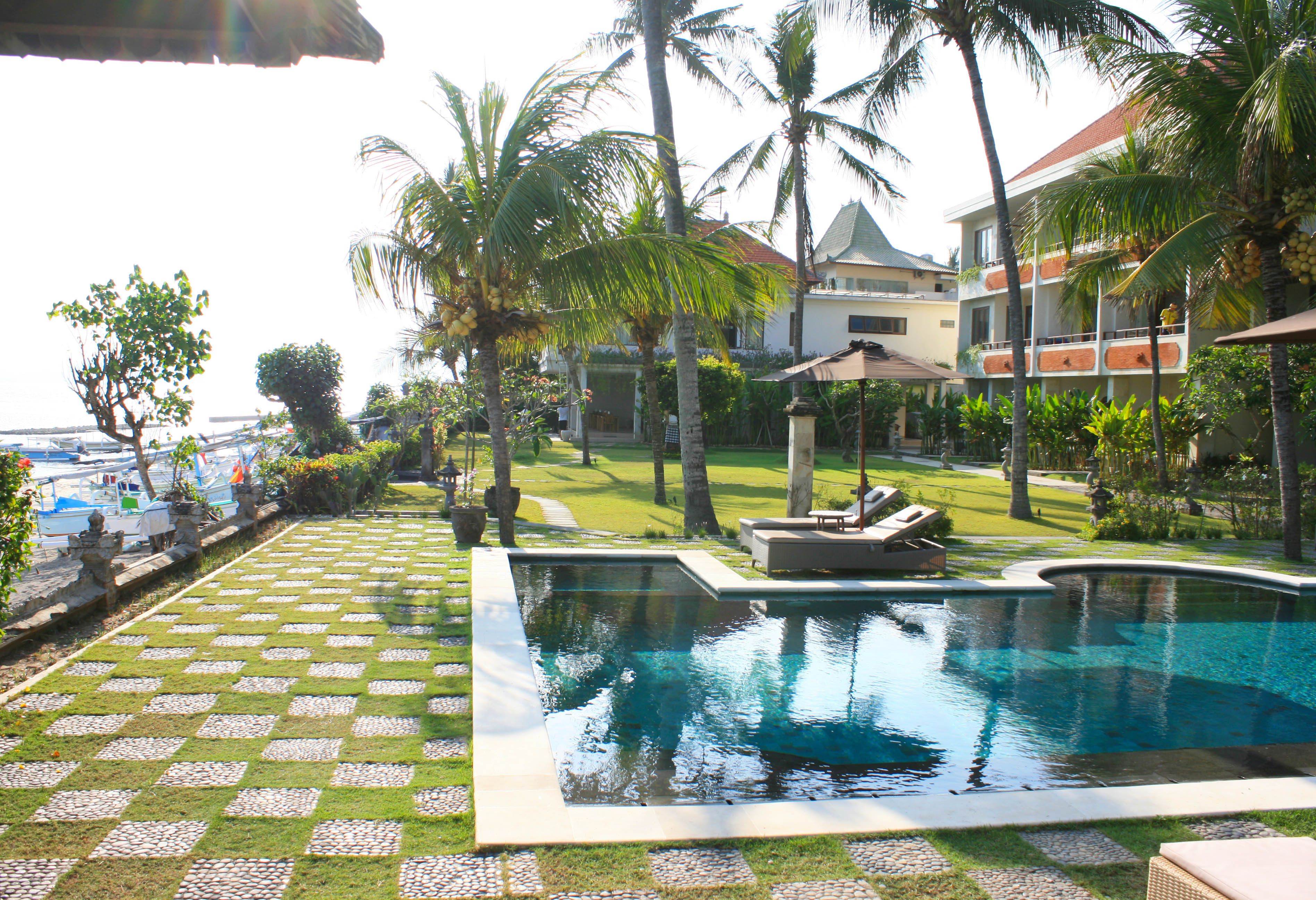 A swimming pool with a checkered pattern of grass and pebbles, surrounded by palm trees and a building with a red roof.