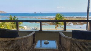Un balcone con mobili in vimini che si affaccia su una spiaggia con acque cristalline e coste rocciose.