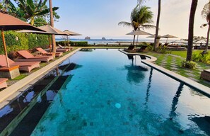 A swimming pool with lounge chairs and umbrellas, surrounded by palm trees and a view of the sea. - Hotel Genggong (Karangasem)