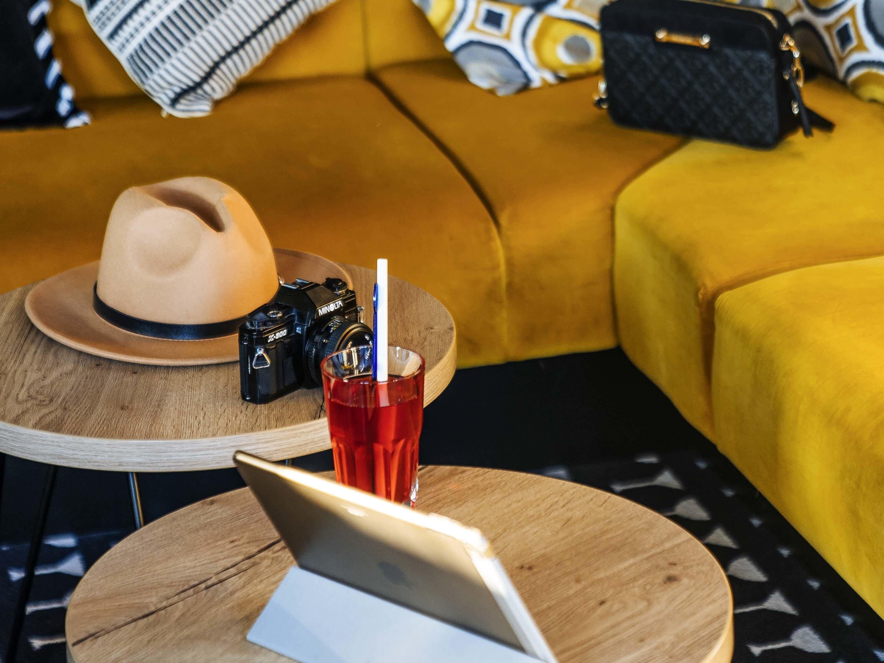 A wooden table with a laptop, a camera, a drink, and a hat.