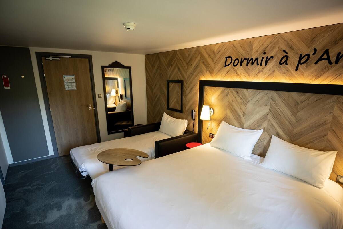 Une chambre d’hôtel avec deux lits, un miroir, une table de chevet avec une lampe et un mur en bois orné de texte.