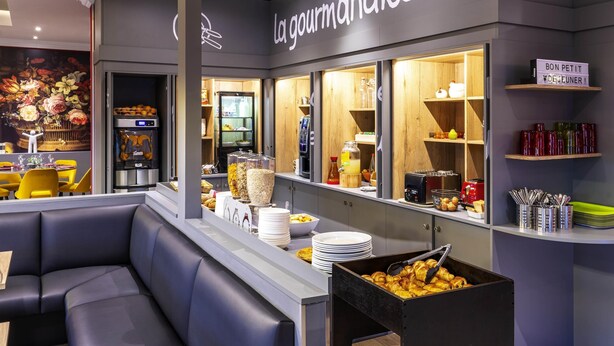 Een modern caféinterieur met zitplaatsen, een toonbank met etenswaren en een bordje met de tekst 'la gourmandise'.