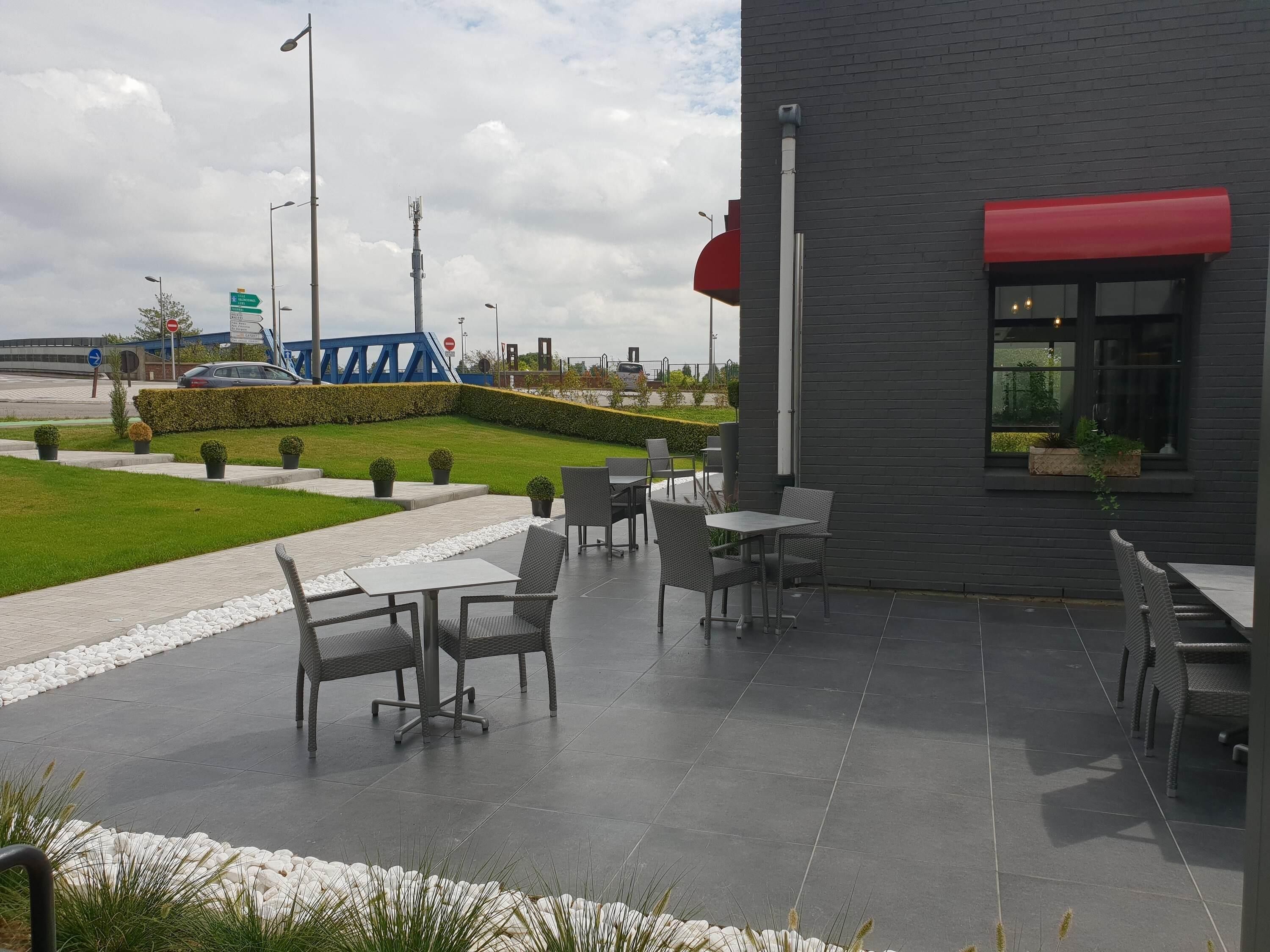 An outdoor patio area with tables and chairs, a hedge, and a building with a red awning.