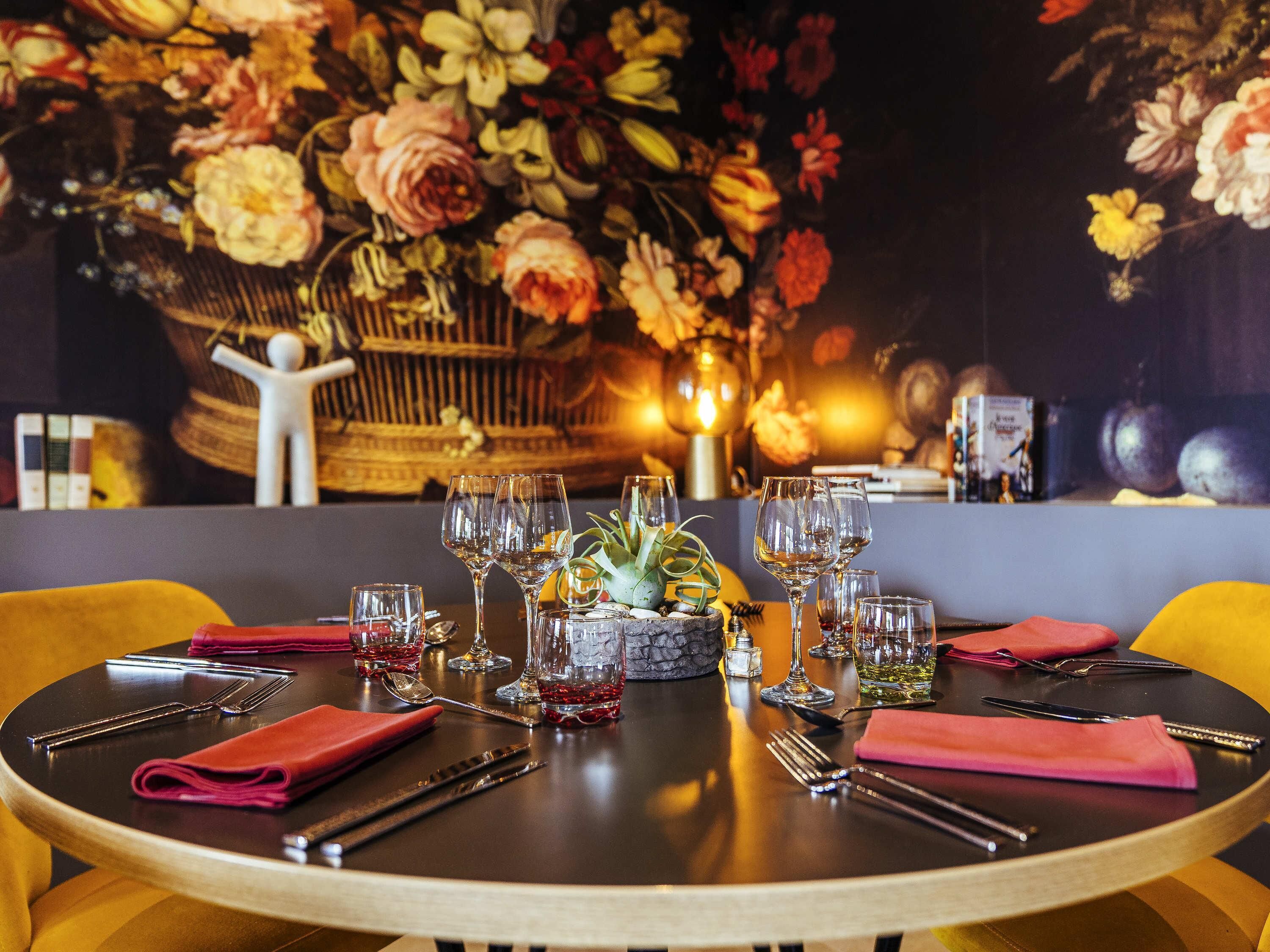 Mesa redonda para comer dispuesta con copas de vino, cubiertos y servilletas, rodeada de coloridas decoraciones florales y una jaula decorativa.