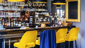 Een bar met verschillende drankflessen, glazen en een koffieapparaat. Er zijn gele stoelen en een blauwe stoel.