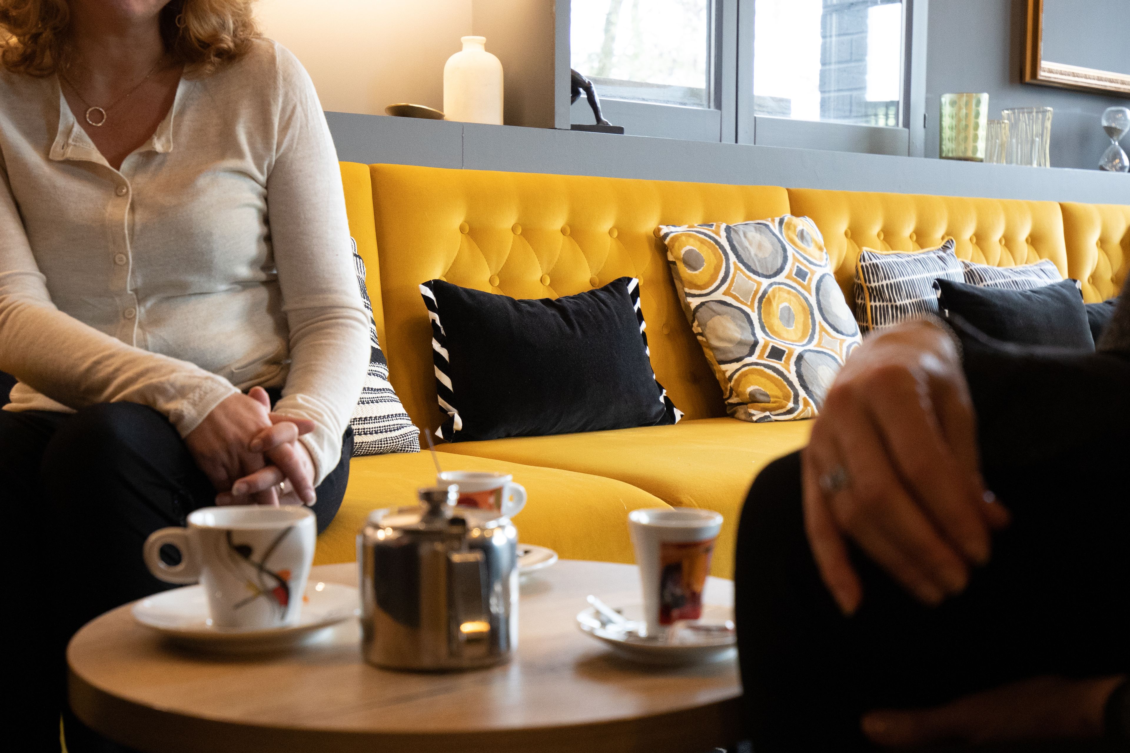 Un canapé jaune agrémenté de coussins noirs et à motifs, une table ronde avec une cafetière et des tasses, et une fenêtre donnant sur des immeubles.