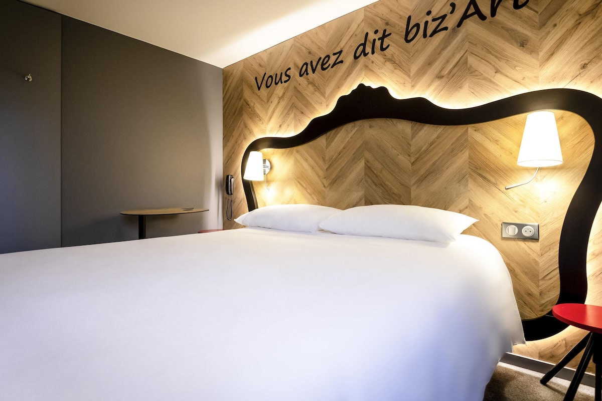 Une chambre d’hôtel avec un lit, une table de chevet, un fauteuil rouge et un mur orné d’un motif en chevrons en bois, ainsi qu’un luminaire.