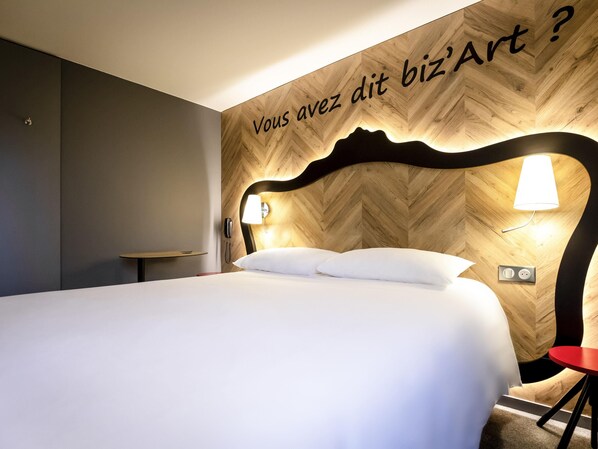 Ein Hotelzimmer mit Bett, Nachttisch, einem roten Sessel sowie einer Wand mit Holzvertäfelung im Fischgrätmuster und einer Leuchte.