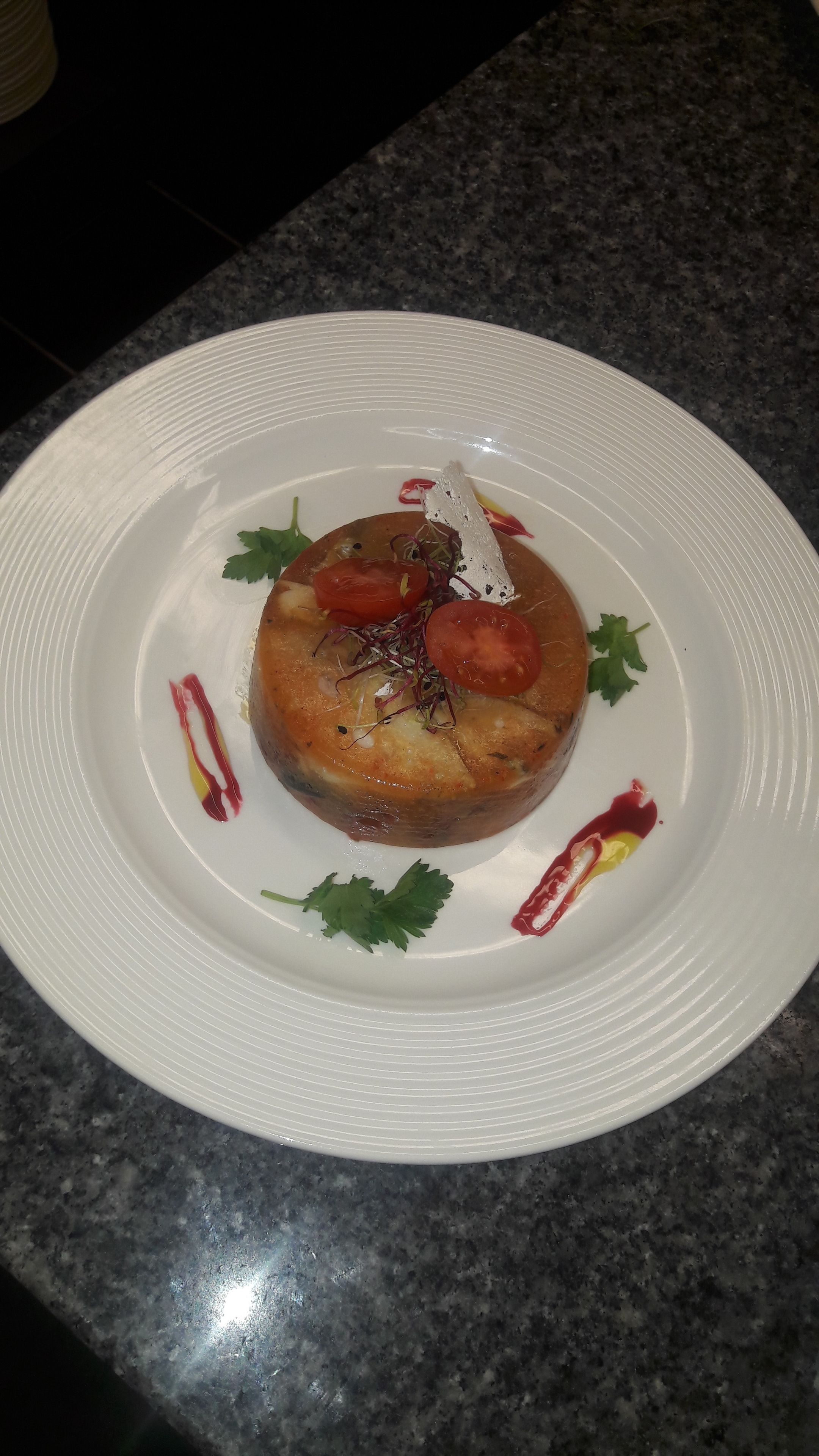 A plate with a round, golden-brown dish, garnished with parsley and tomato slices, on a white plate with a textured rim.