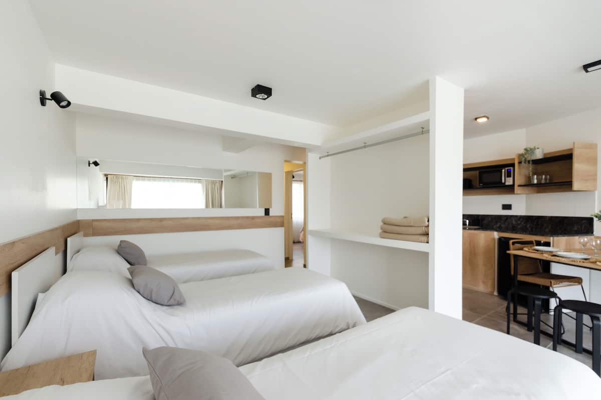 A modern hotel room with two beds, a small kitchenette, and a dining area.