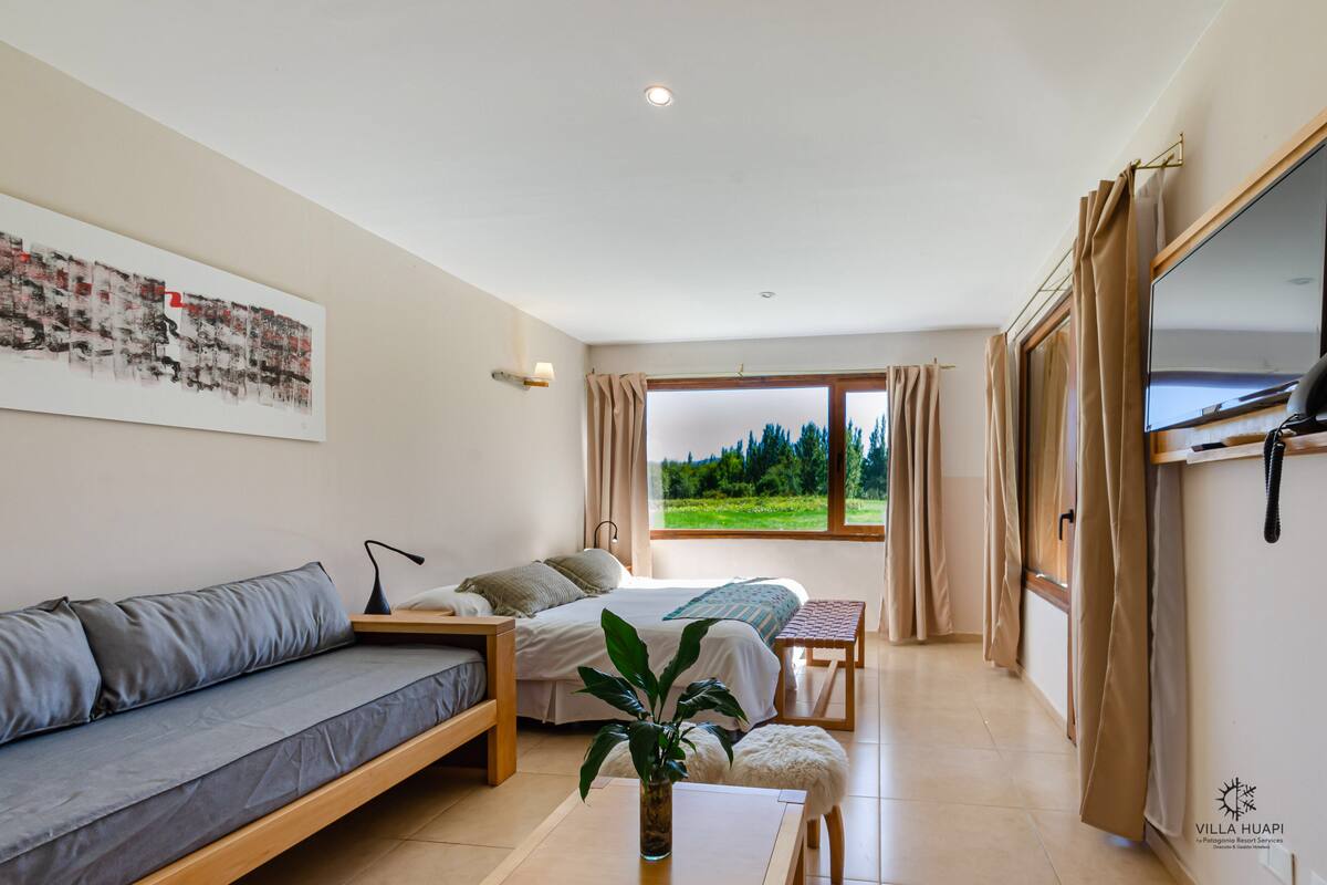 A modern bedroom with a bed, a sofa, a desk, and a large window offering a view of greenery.
