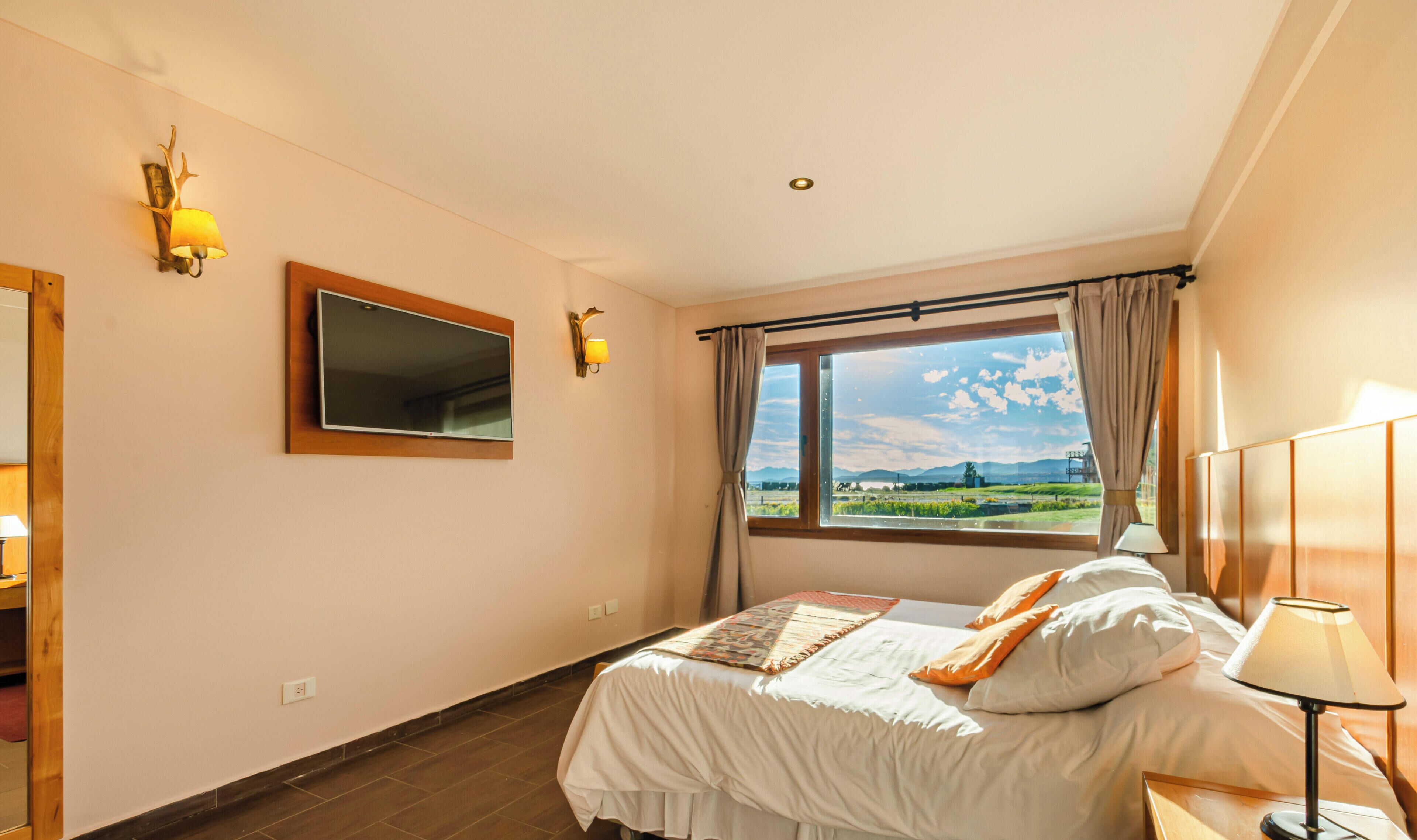 Quarto de hotel com cama, janela com vista, espelho e luzes fixadas na parede.