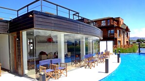 A modern hotel with a glass-enclosed pool area, outdoor seating, and a view of the mountains. - Villa Huapi (Dina Huapi)