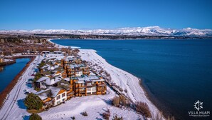 A snow-covered waterfront property with multiple buildings, a clear view of the lake, and mountains in the background. - Villa Huapi (Dina Huapi)