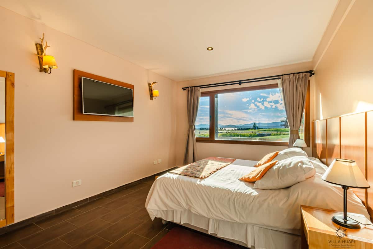A hotel room with a bed, a window with a view, a mirror, and wall-mounted lights.