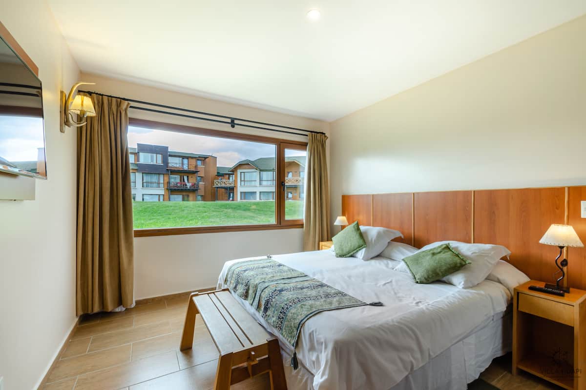 A hotel room with a large bed, a bench, a nightstand, and a view of a building and lawn outside.