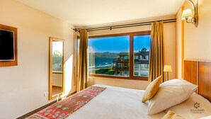A hotel room with a large window offering a view of a coastal area, a bed with pillows, a bedside table with a lamp, and a mounted TV. - Villa Huapi (Dina Huapi)