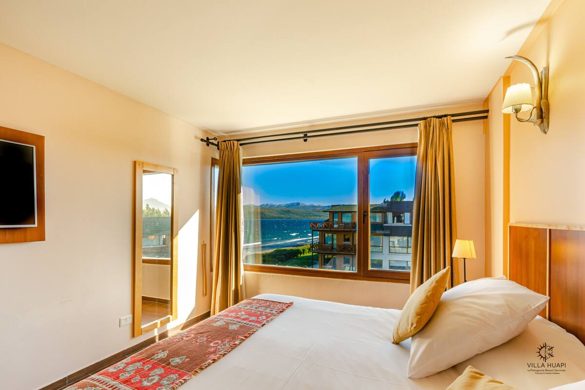 A hotel room with a large window offering a view of a coastal area, a bed with pillows, a bedside table with a lamp, and a mounted TV.