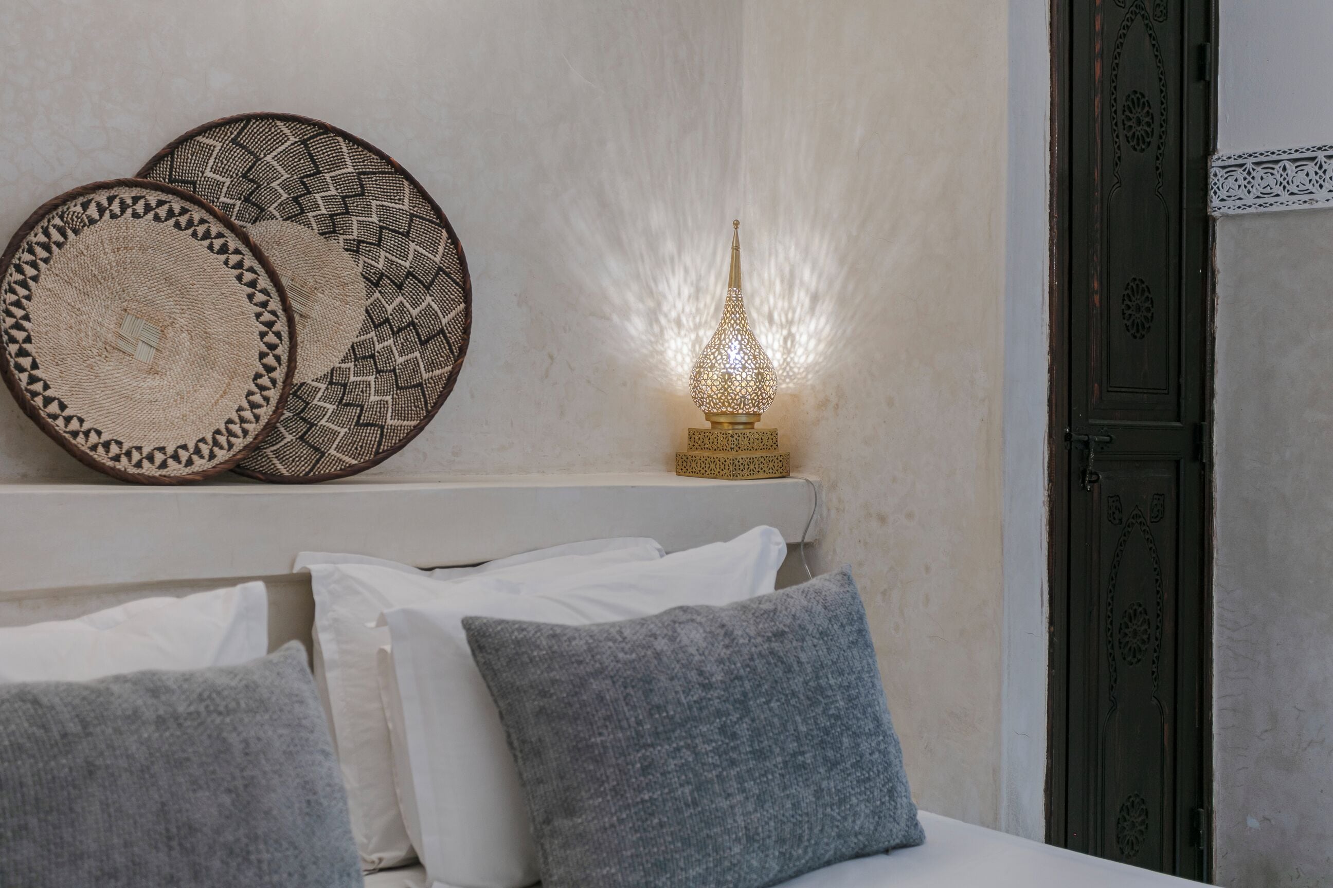A neatly arranged bed with white and gray pillows, a decorative wall hanging, and a wall-mounted lamp.