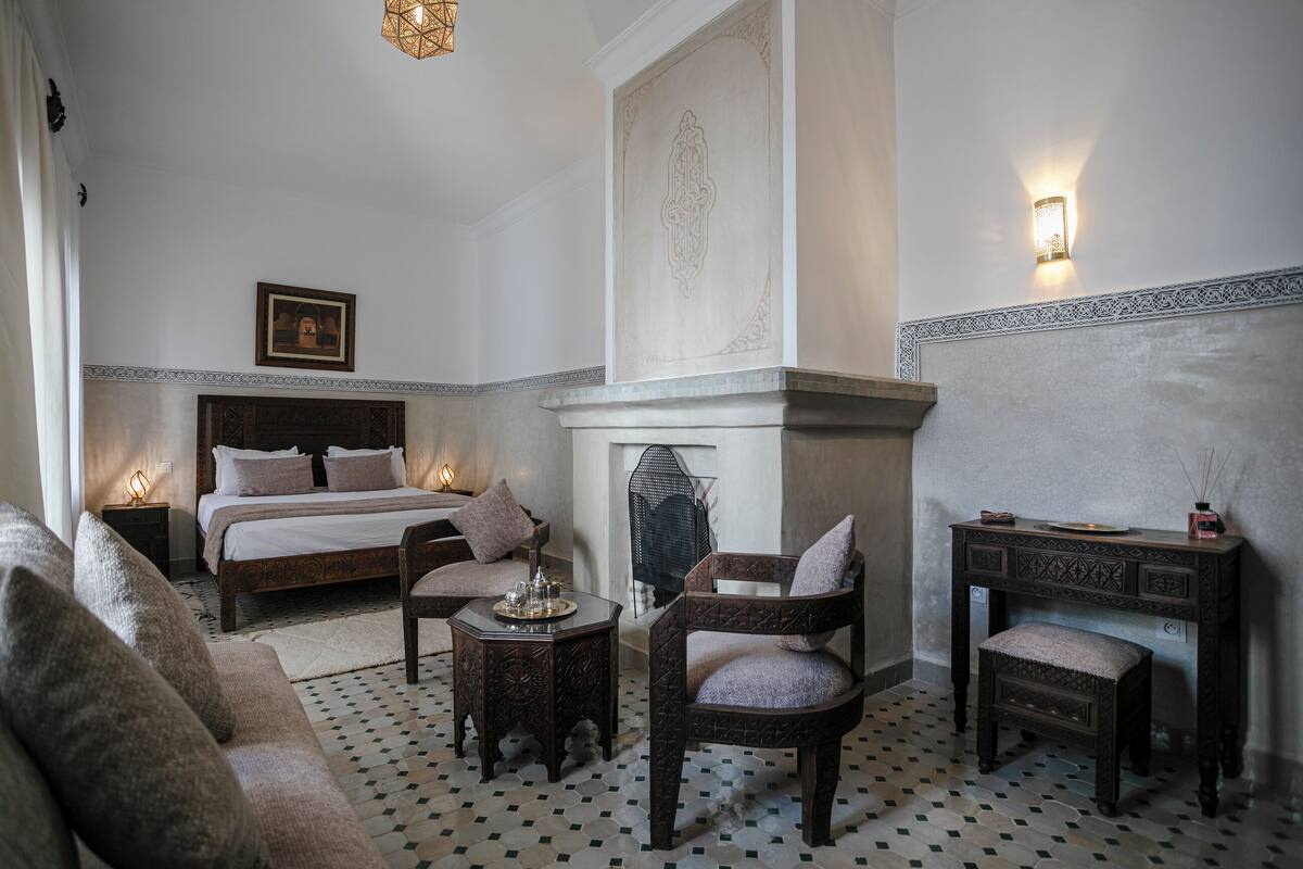 A bedroom with a bed, a chair, a small table, a nightstand, a fireplace, and a wall-mounted light fixture.