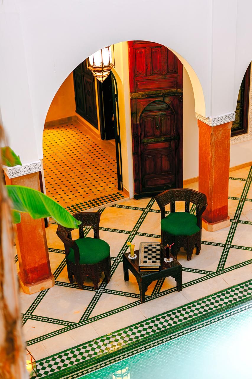 A room with a checkered floor, two chairs with green cushions, a small table with a chessboard, and a door with a red frame.