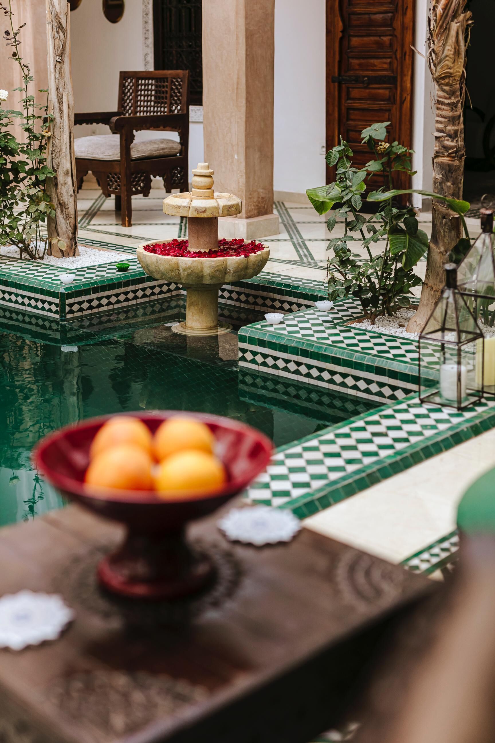 A pool area with a fountain, a chair, and a table with fruit.