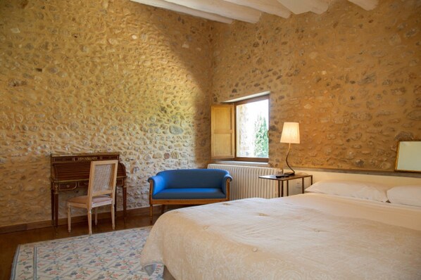 Un dormitorio con pared de piedra, una cama, un sillón azul, un escritorio de madera y una lámpara.