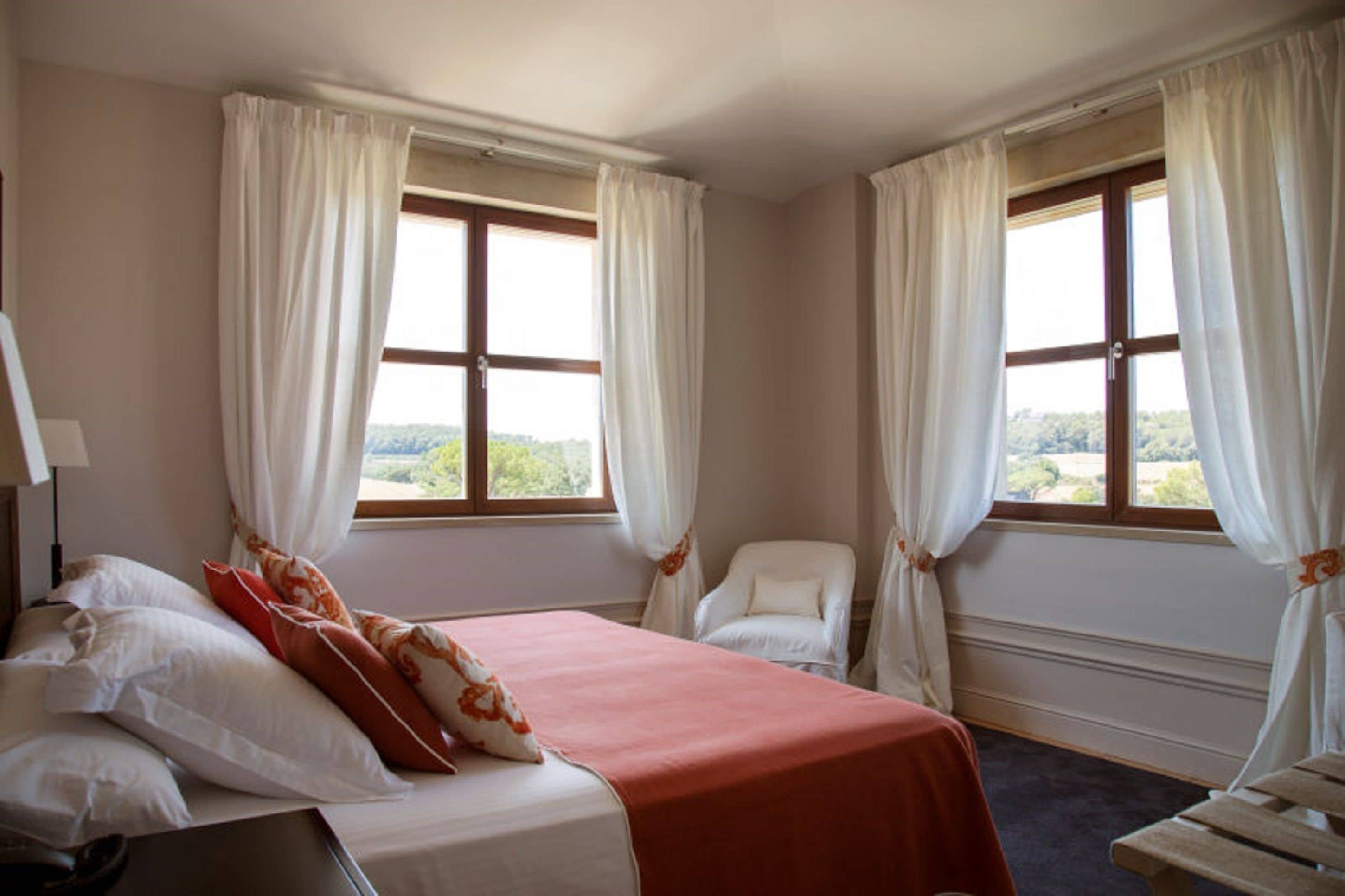 Una habitación de hotel con cama, dos ventanas con cortinas, una silla y un escritorio con lámpara.