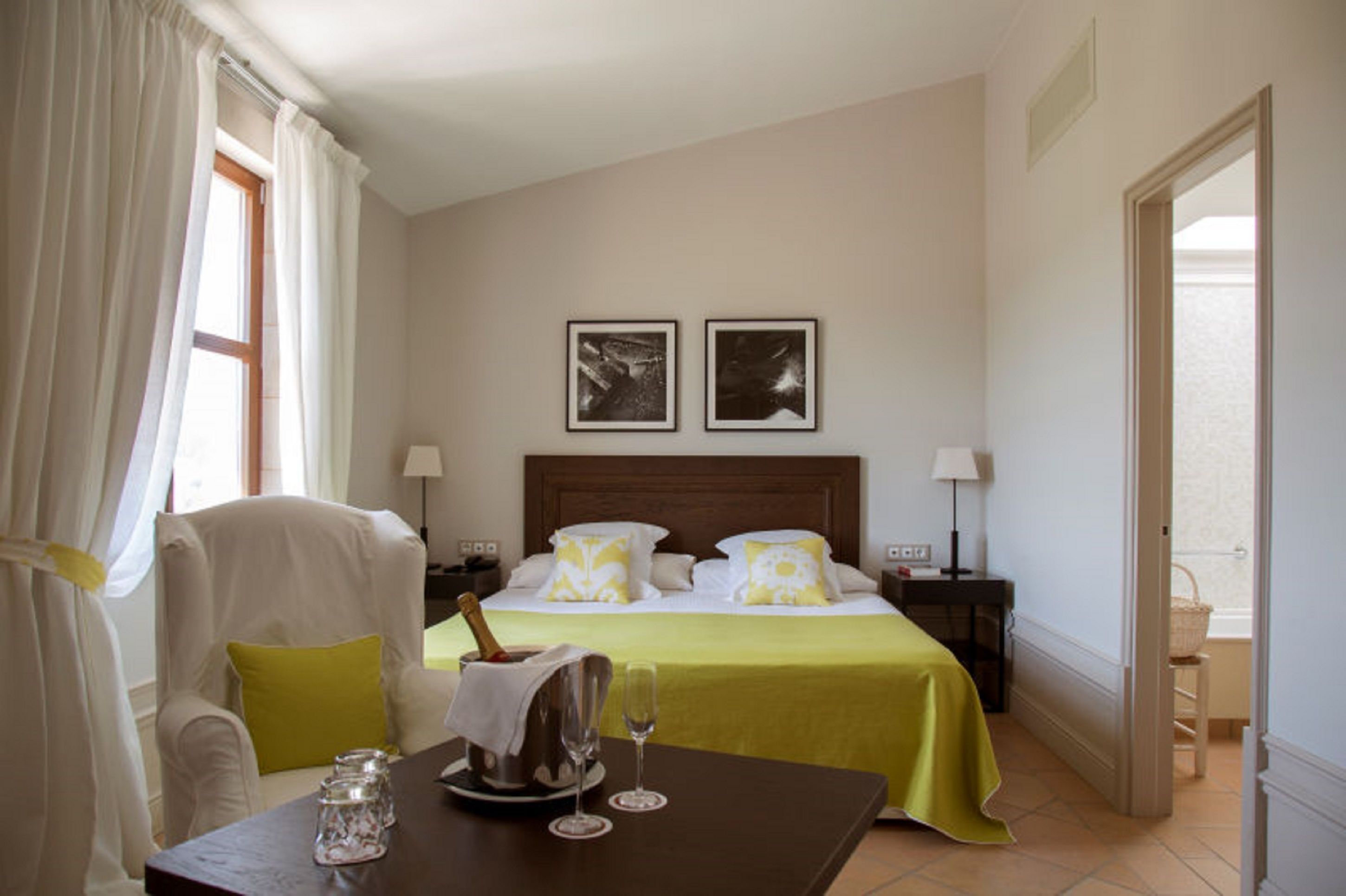 Quarto de hotel com cama, uma mesa com uma garrafa de champanhe, duas quadros na parede e uma cadeira.