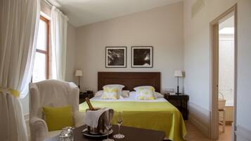 Quarto de hotel com cama, uma mesa com uma garrafa de champanhe, duas quadros na parede e uma cadeira.