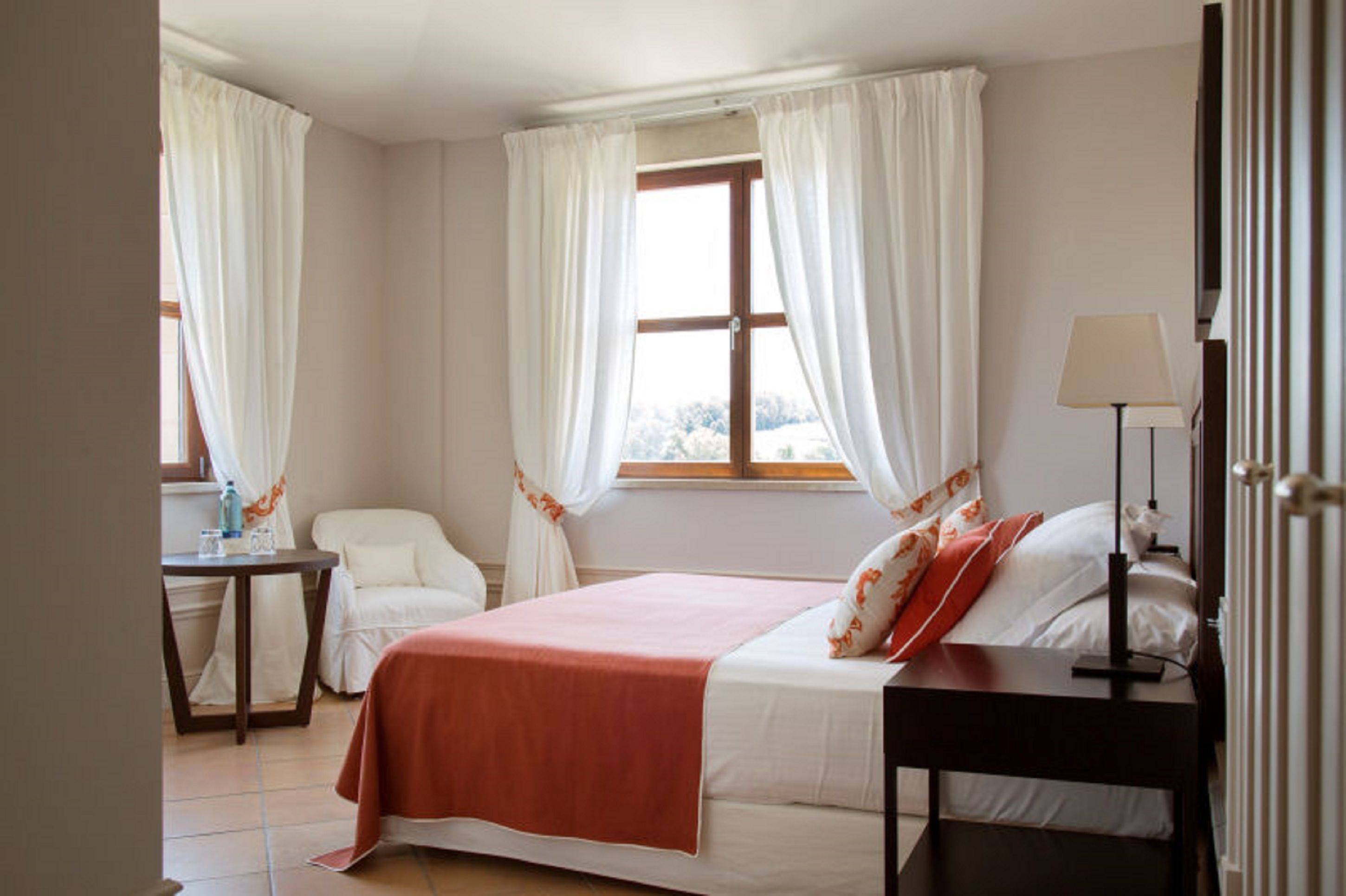 A bedroom with a bed, a bedside table, a chair, a window with curtains, and a lamp.