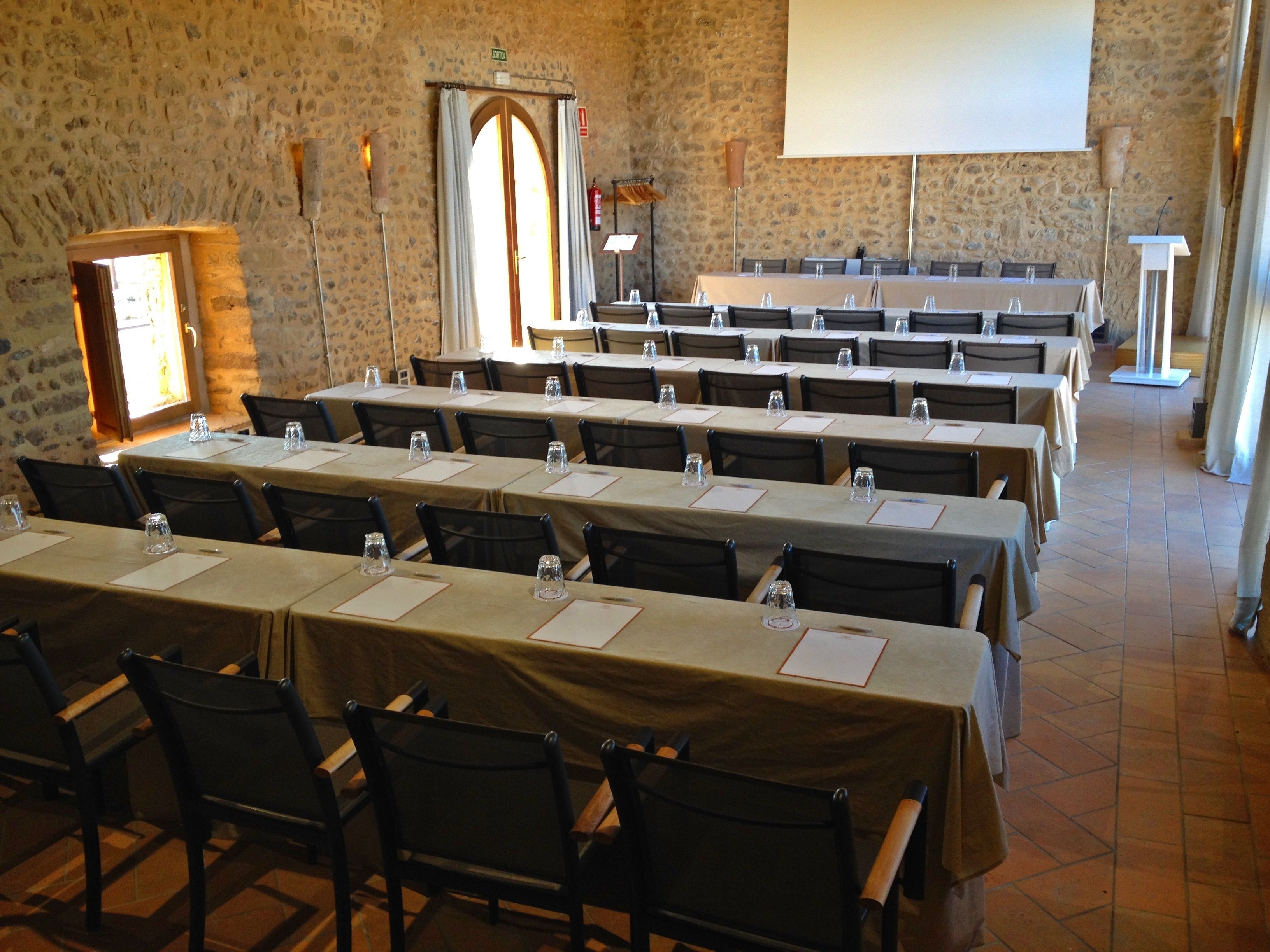 A conference room with multiple tables and chairs, stone walls, a projector screen, and a podium.
