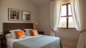 A bedroom with a large bed, two framed pictures on the wall, a nightstand with a lamp, and a window with curtains.