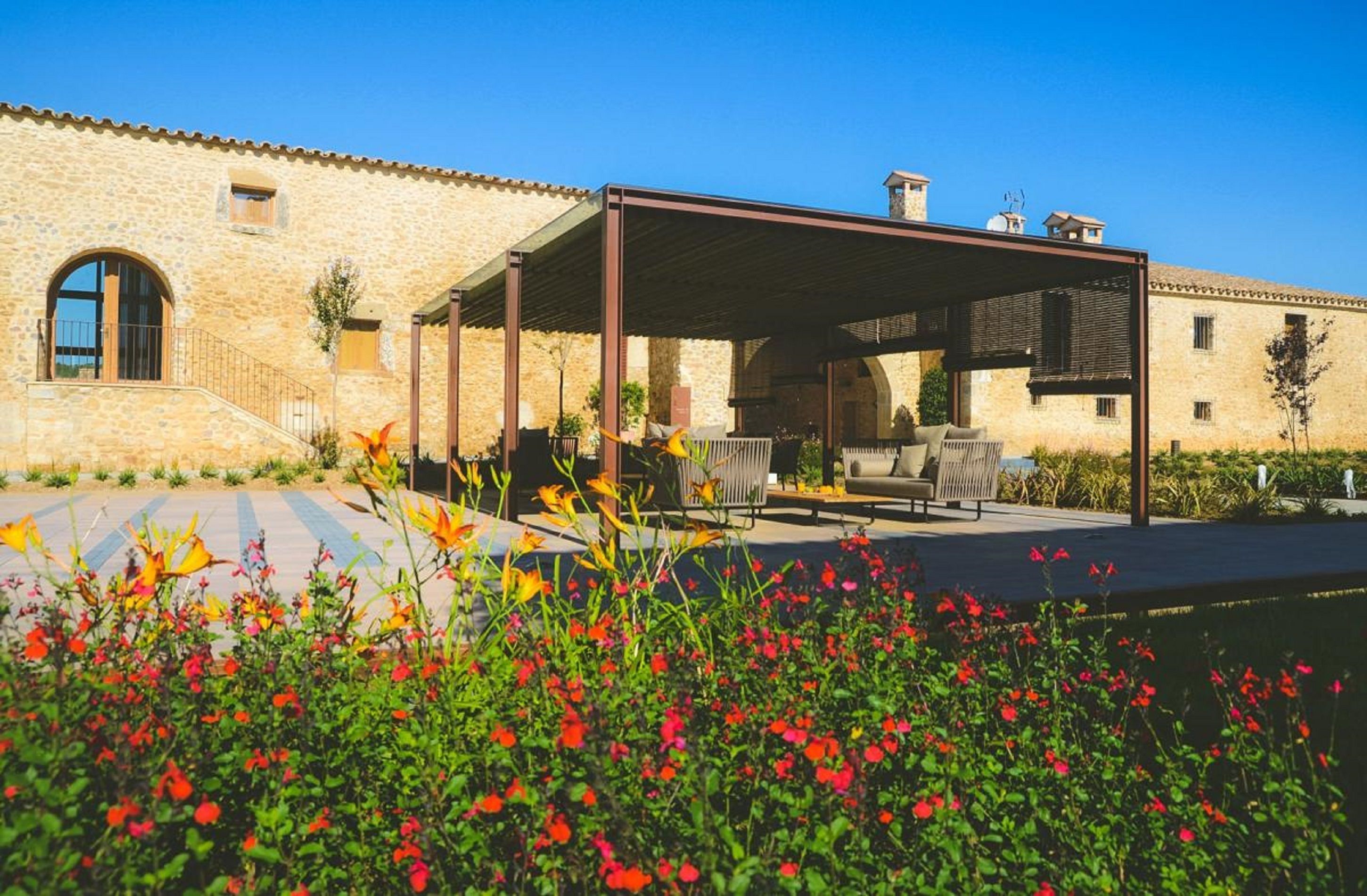 Un bâtiment en pierre doté d’un patio couvert, d’un espace extérieur aménagé pour s’asseoir et d’un jardin avec des fleurs rouges.