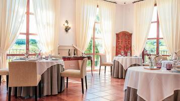 A dining area with tables set for two, large windows with curtains, and a view of greenery outside.