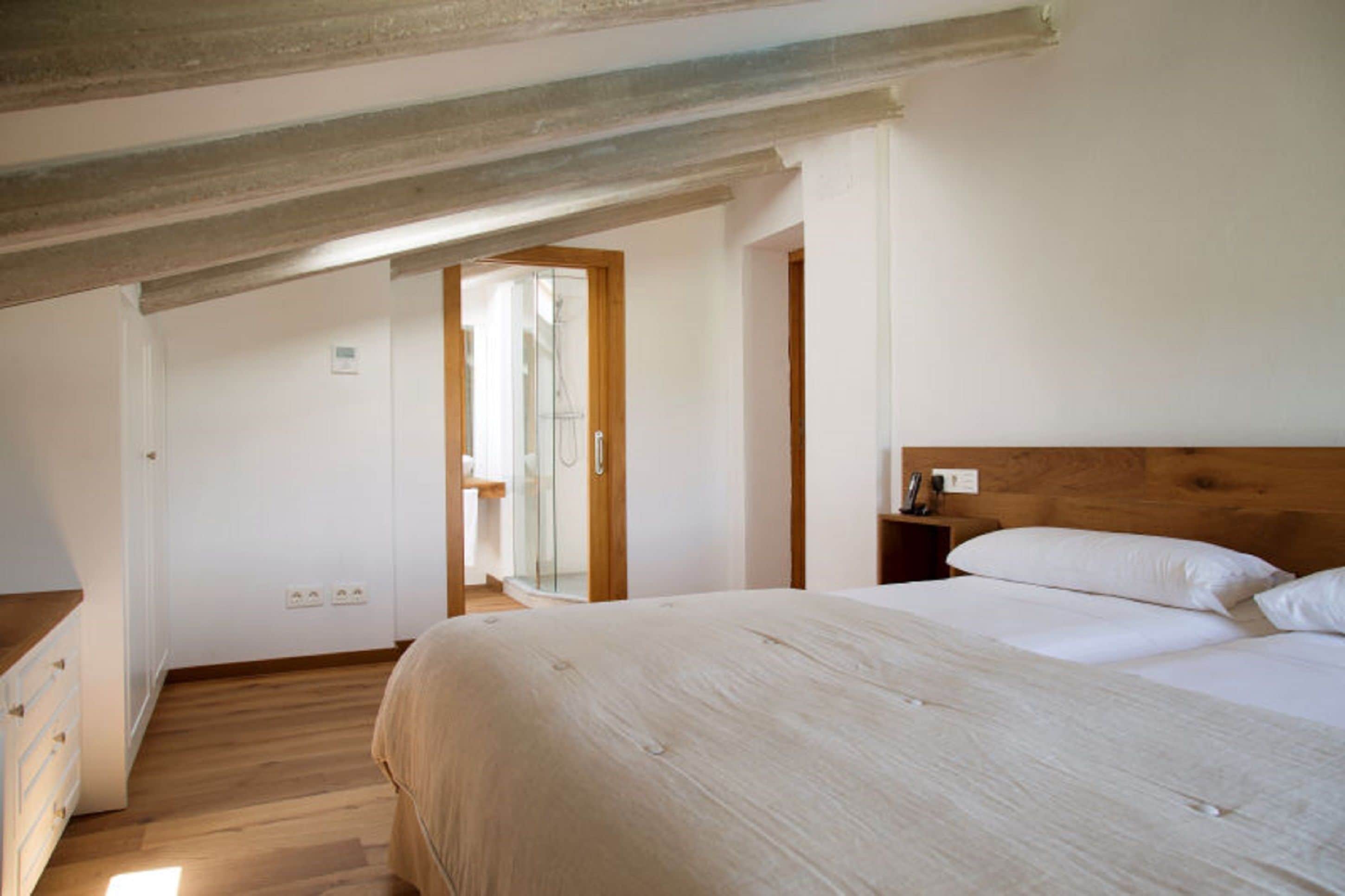 A bedroom with a wooden ceiling, a bed with white bedding, a nightstand, and a window with a view of the outside.