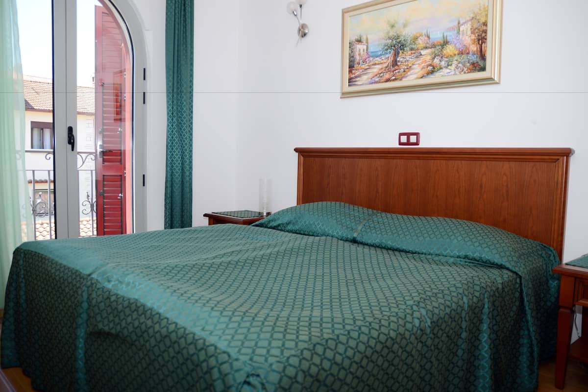 Ein Schlafzimmer mit hölzernem Kopfende, einer grün gemusterten Tagesdecke, einem Bild an der Wand und Blick durch eine Glastür.