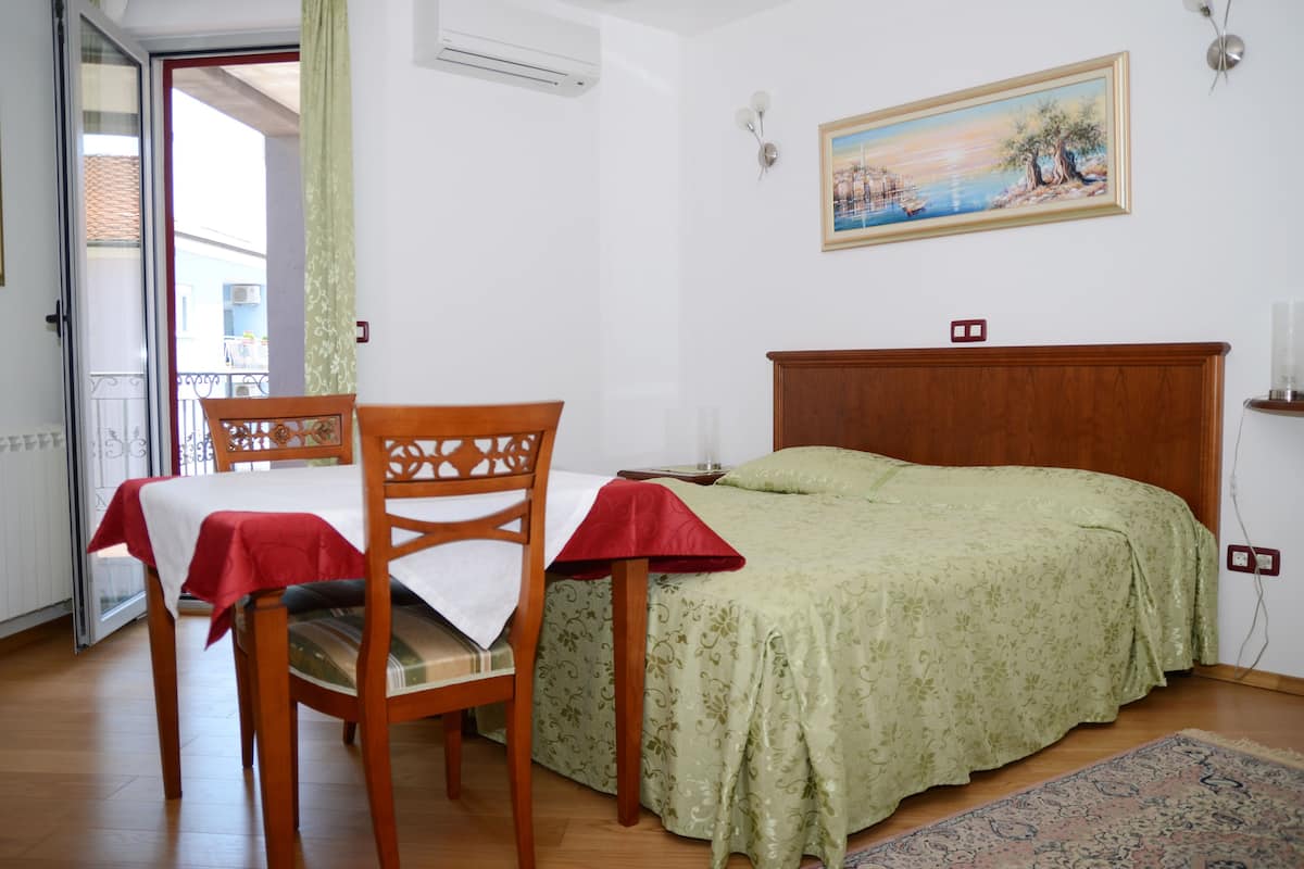 Ein Schlafzimmer mit Bett, Sessel, Tisch mit rotem Tuch und einem Bild an der Wand, sowie Blick durch eine offene Tür.