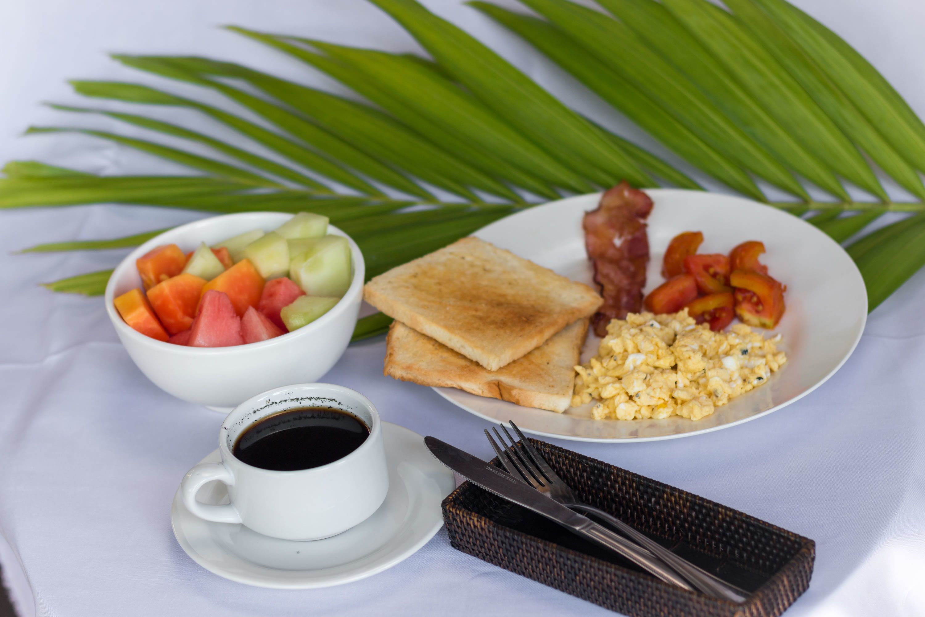 A breakfast plate with toast, scrambled eggs, bacon, and a bowl of fruit.