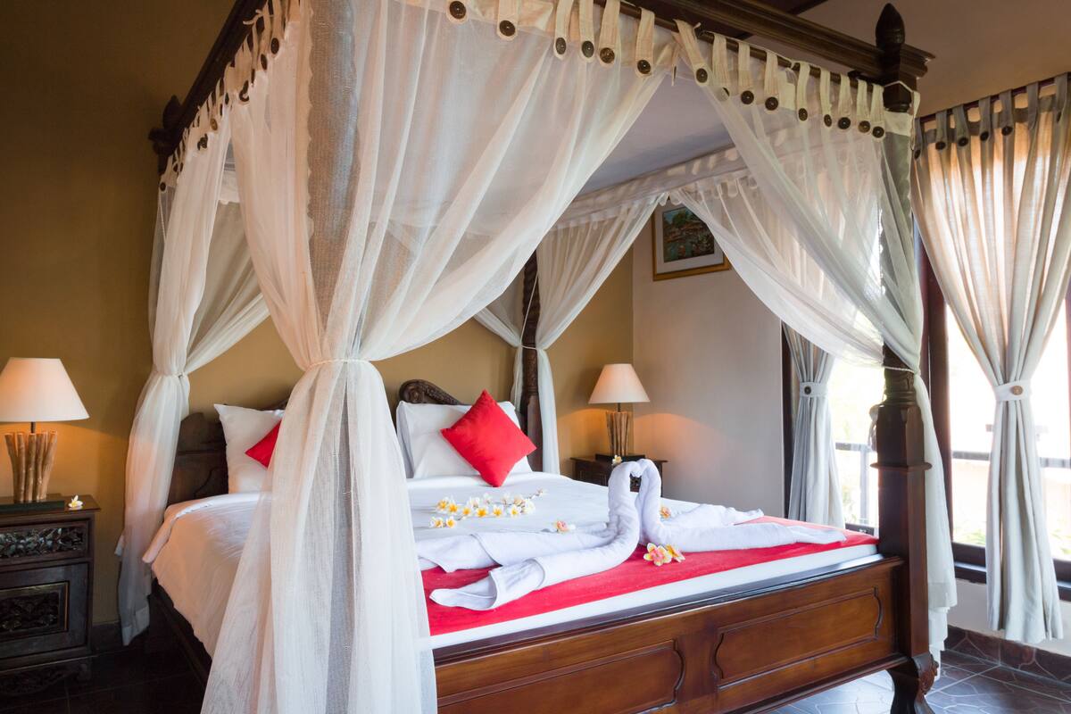 Una cama con dosel y cuatro columnas con una colcha roja y blanca, almohadas blancas y una decoración de toallas con forma de cisne.