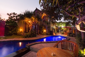 A swimming pool with a curved edge, surrounded by tropical plants and a wooden deck. - Pondok Mimpi Tulamben (Tulamben)
