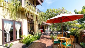 Ein Innenhof mit Tisch und Stühlen unter einem großen Sonnenschirm, umgeben von Kübelpflanzen und einem gepflasterten Weg.