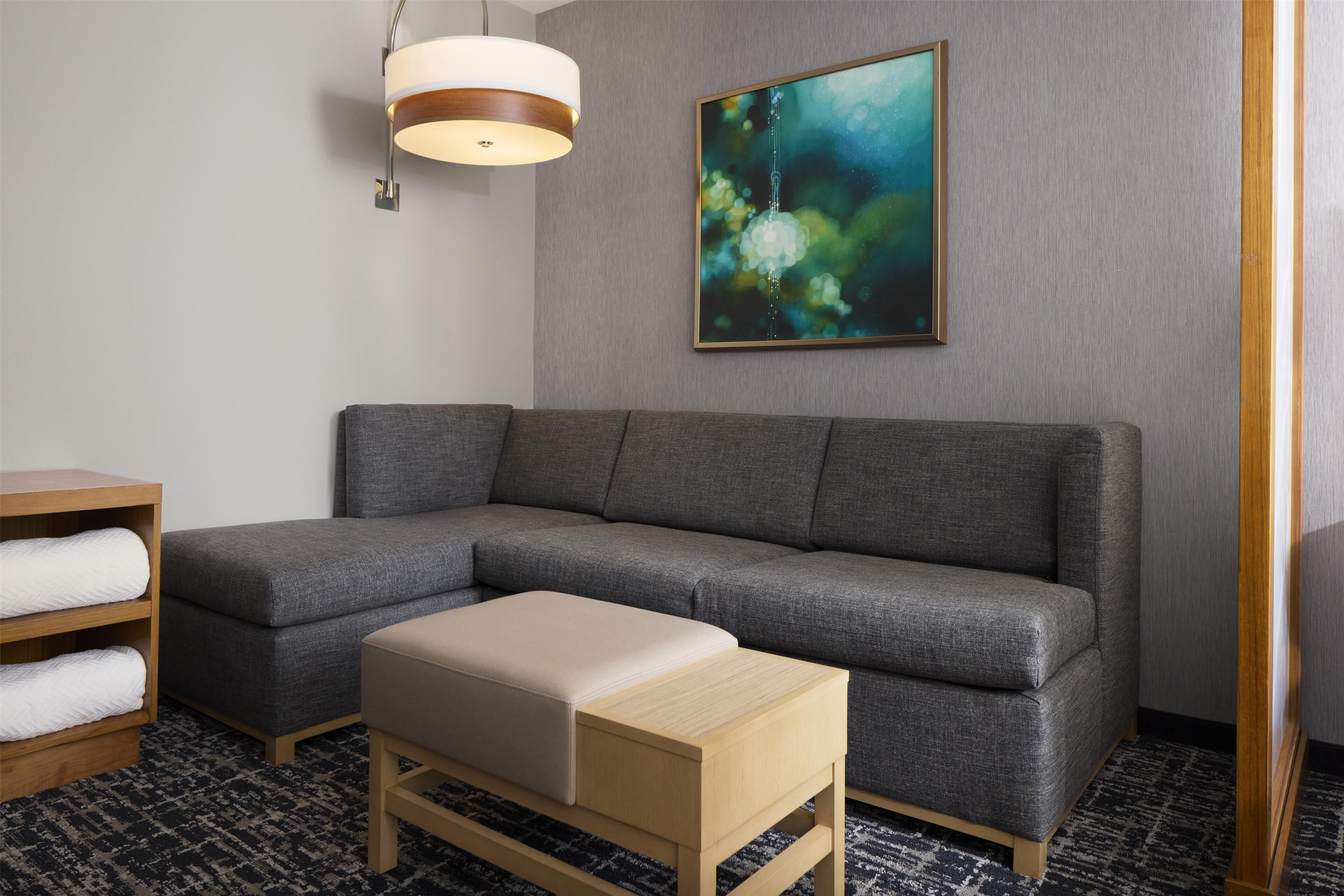 A modern hotel room with a grey sectional sofa, a wooden side table, a beige ottoman, a wall-mounted abstract painting, and a hanging pendant light.