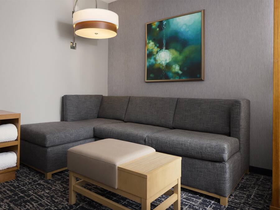 Ett modernt hotellrum med en grå, modulbaserad soffa, ett träbord, en beige pall, en abstrakt tavla som hänger på väggen och en hängande taklampa.