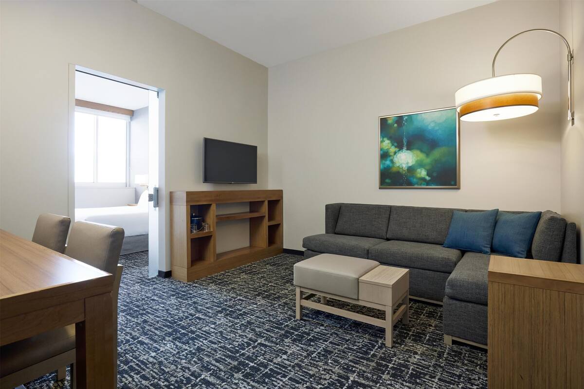 A modern hotel room with a grey sofa, a wooden coffee table, a flat-screen TV, a bookshelf, and a large abstract painting.