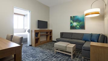 Een moderne hotelkamer met een grijze bank, een houten salontafel, een flatscreen televisie, een boekenkast en een groot abstract schilderij.