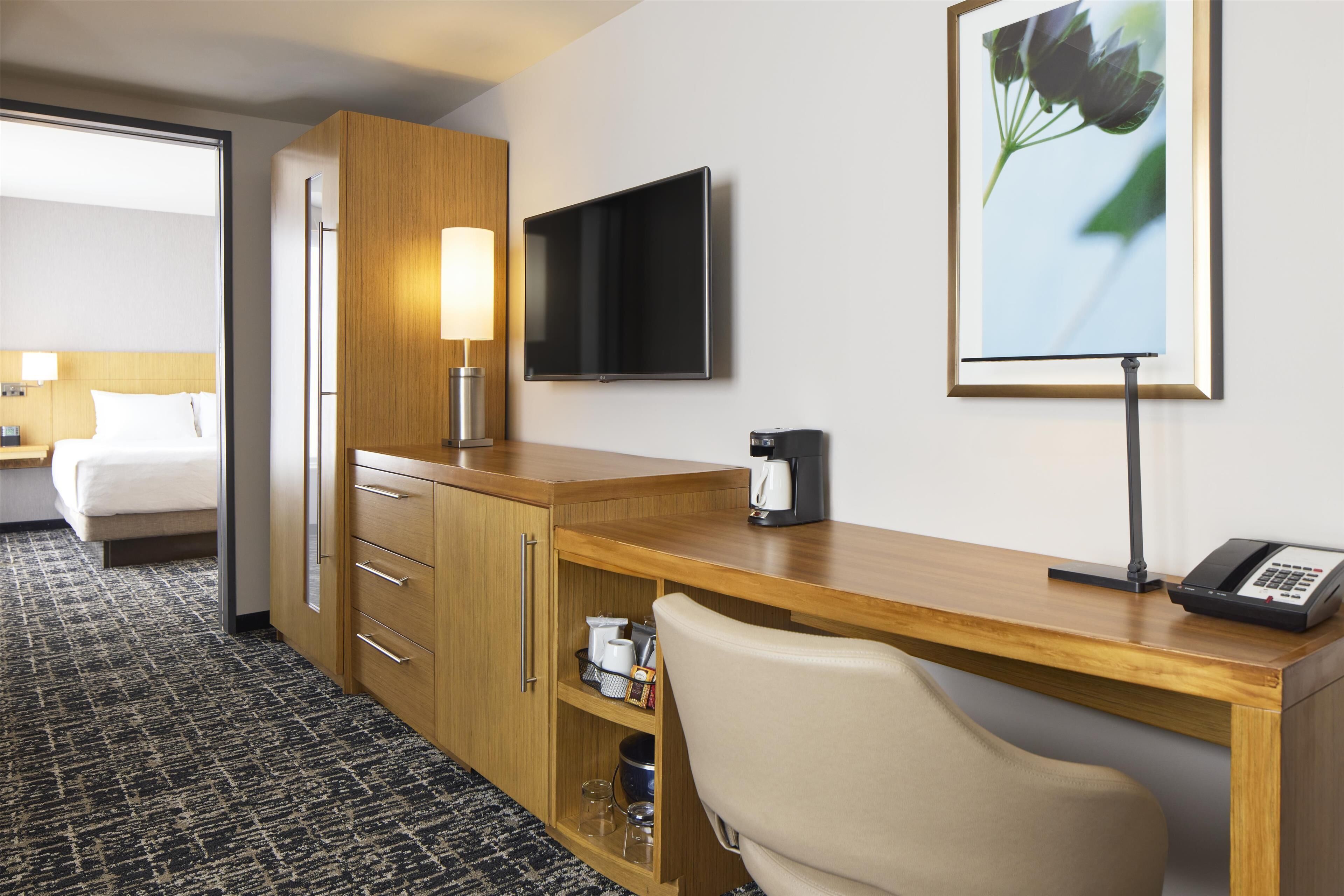 Habitación de hotel con escritorio de madera, televisión de pantalla plana, cafetera, lámpara, silla, cama y un cuadro en la pared.