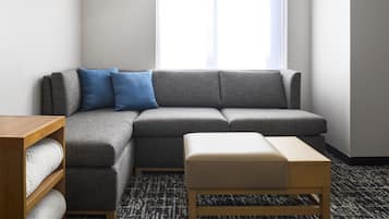 A modern living room with a grey sectional sofa, a wooden side table, a white ottoman, a patterned rug, and a large window with a view.