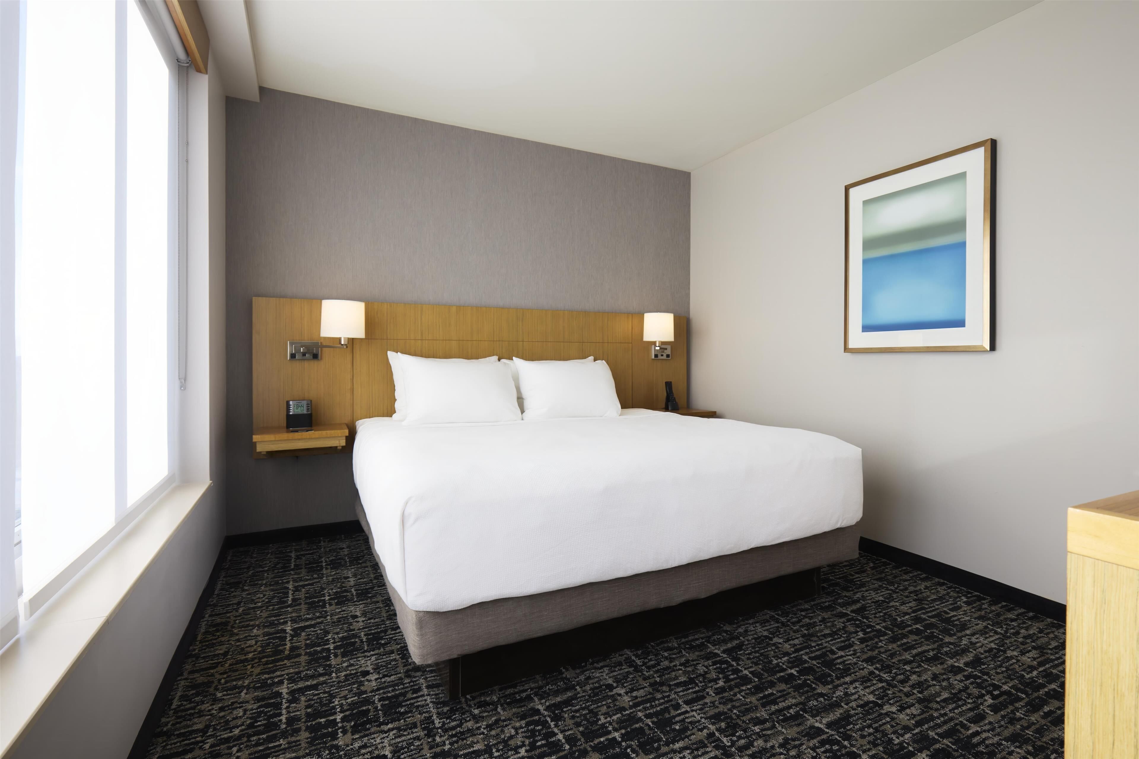 A hotel room with a large bed, two bedside tables with lamps, a nightstand, a framed picture on the wall, and a window providing natural light.