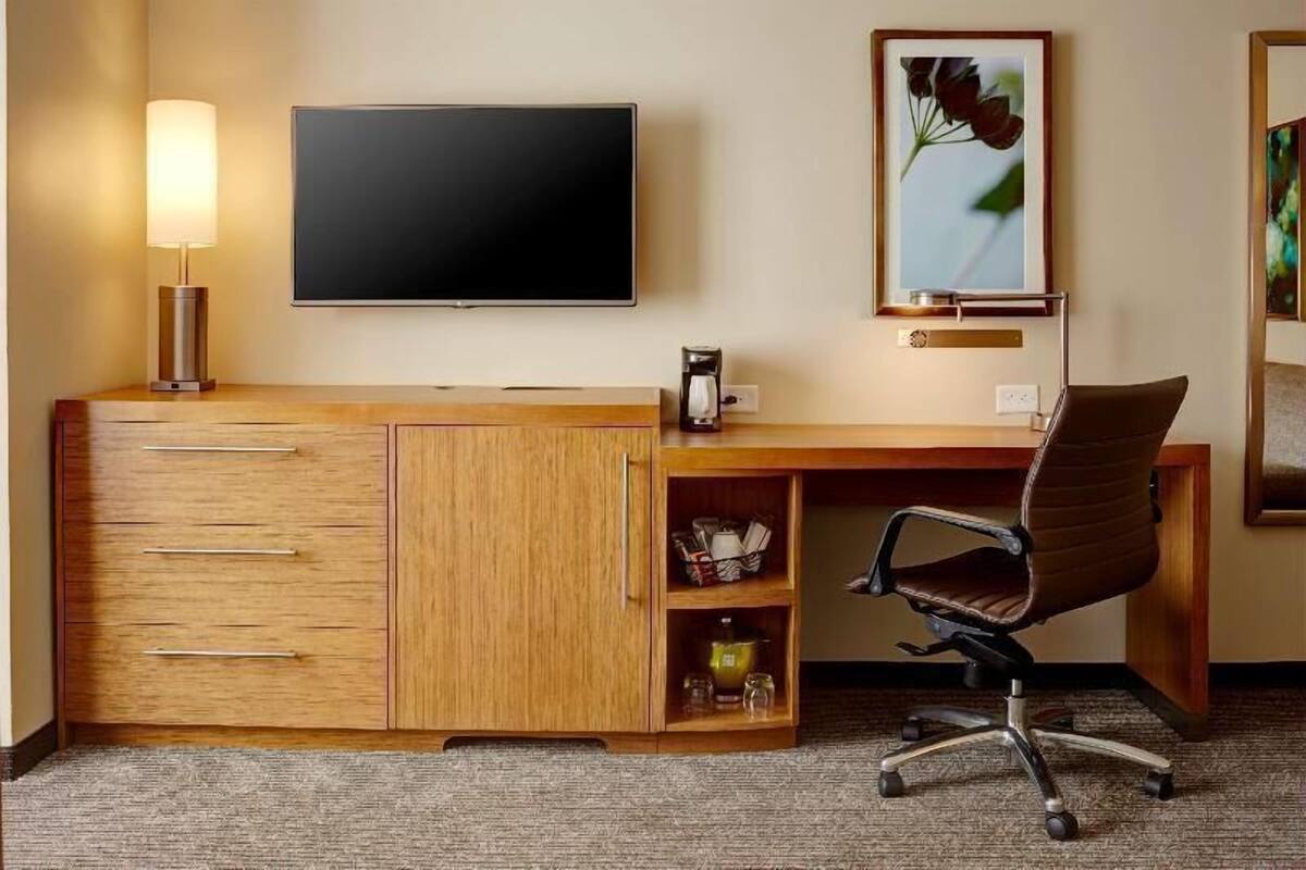 A hotel room with a wooden desk, a flat-screen TV, a coffee maker, a framed picture, and a brown office chair.